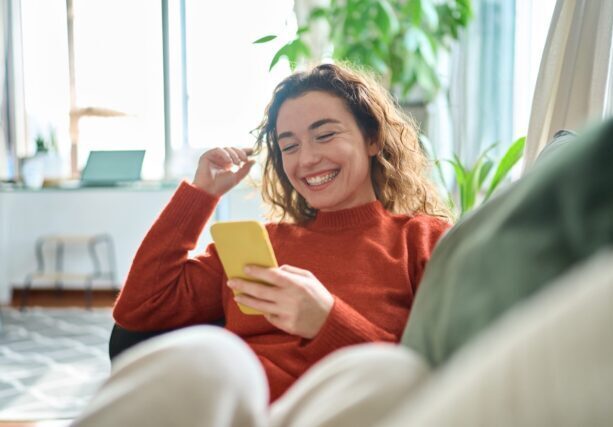 Happy,Relaxed,Young,Woman,Sitting,On,Couch,Using,Cell,Phone,