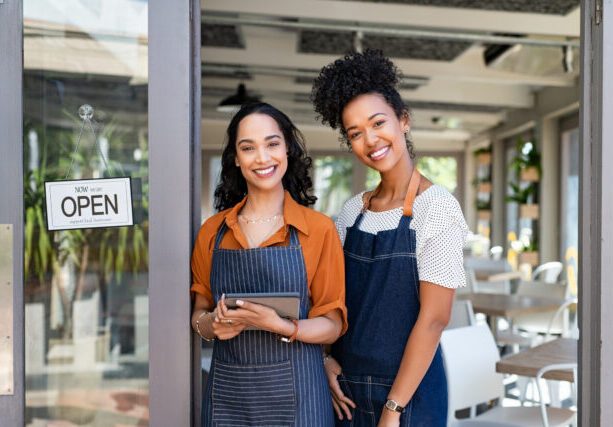 Portrait of smiling young latin woman holding digital tablet with black colleague at cafe entrance door. Two small business owners standing together at cafe entrance while smiling. Happy successful multiethnic small business women wearing apron and standing with open sign at entrance gate of restaurant.