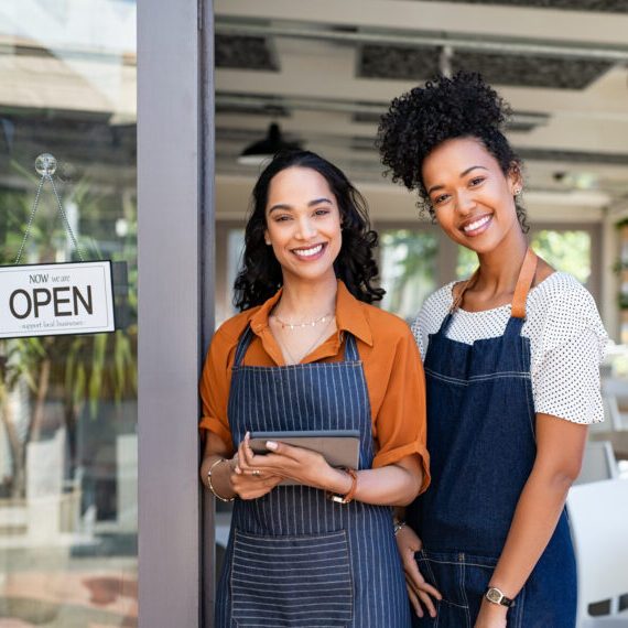 Portrait of smiling young latin woman holding digital tablet with black colleague at cafe entrance door. Two small business owners standing together at cafe entrance while smiling. Happy successful multiethnic small business women wearing apron and standing with open sign at entrance gate of restaurant.